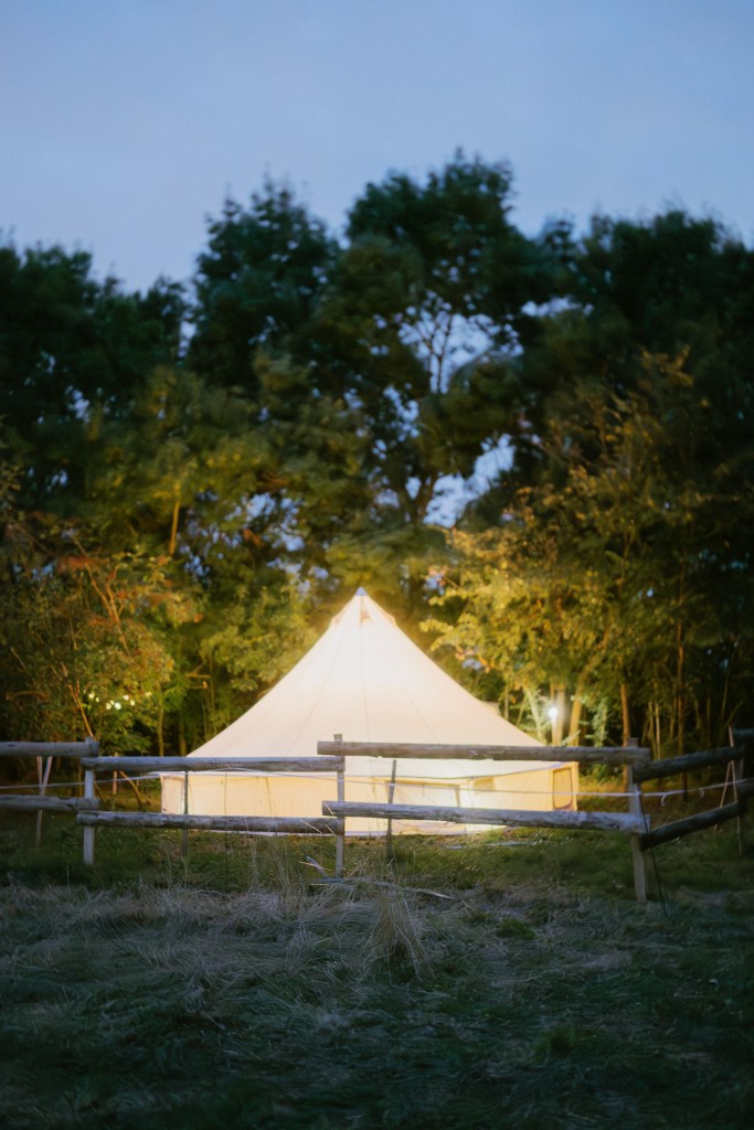 A small white tent is pitched in a field, surrounded by trees, a wooden fence, and lit from within, set against a bluish sky.